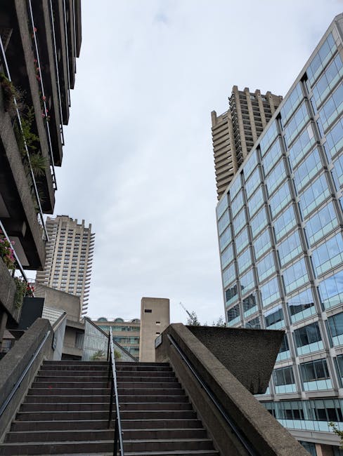 A multi-storey residential building in an urban setting, featuring rows of balconies with metal railings and wooden window frames. The balconies have various potted plants and greenery, some cascading